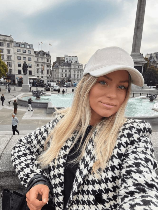 En profilbild på Julia Bergman med en vit keps och en svart-vit-rutig jacka. Hon ler in i kameran och fotar en selfie framför en fontän.