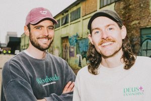 En bild på duon Tim och Danny från ArgaSkånskaMän. De ler in mot kameran och medan Tim har på sig en rosa keps och Danny har på sig en grön keps. Tim håller armarna i kors i en grå tröja med tryck på och Danny har en vit T-shirt med tryck på.