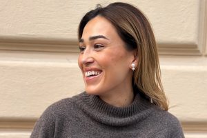 En bild på Vanessa Valderrama som tittar åt sidan och ler. Hon står i en mörkgrå tröja framför en beige vägg.